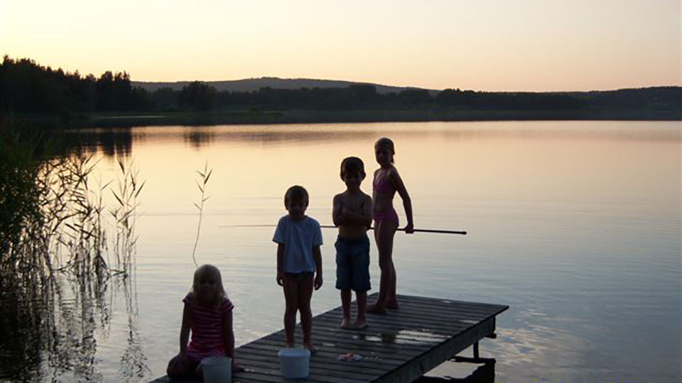 Att fiska är både roligt och spännande för alla. På bryggan sitter och står tre barn och tittar på sin kompis som metar. Sjön Summeln ligger i västra delen av Kila socken i Säffle kommun.