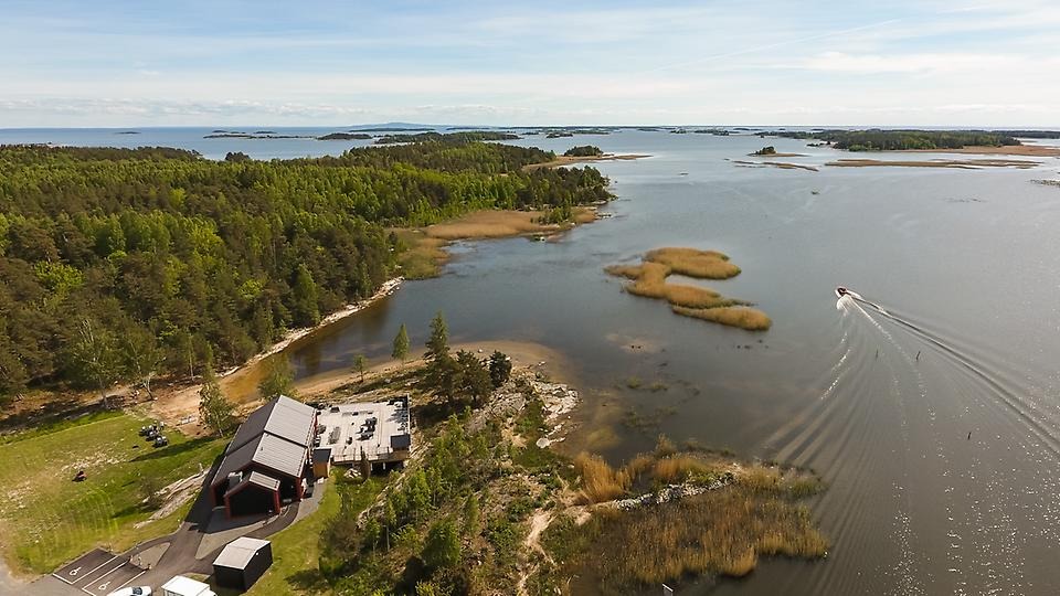 Flygfotot över vackra Ekenäs med camping och restaurang samt över Vänerns vatten. Det är en solig dag och vattnet glittrar, en båt är på väg ut på Vänern.  