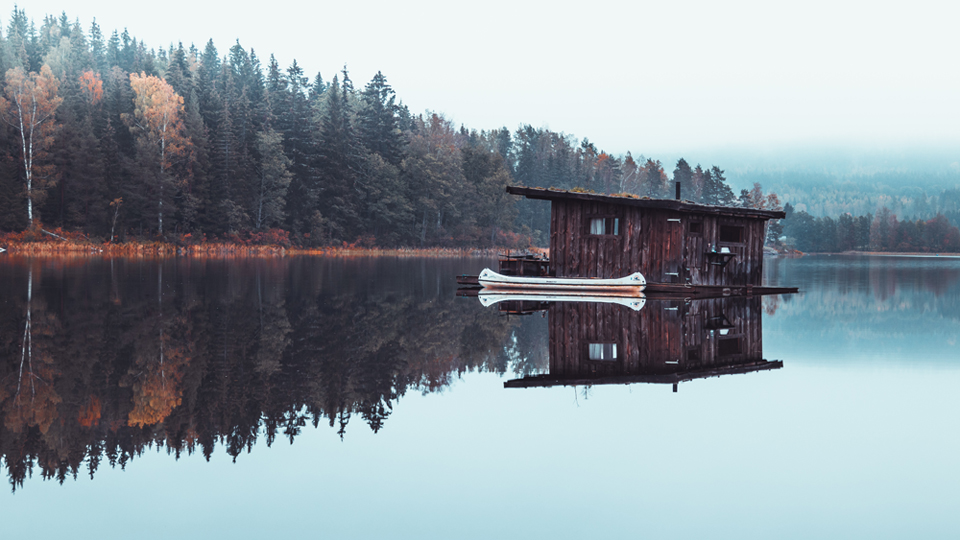 I Naturbyn kan du bo i olika stugor som denna timrade husbåt som ligger i den spegelblanka sjön med en kanot intill husbåten.