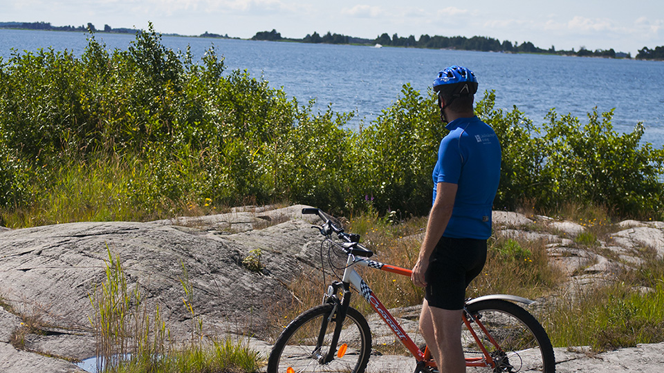 Här står en cyklist med sin cykel på berghällar och ser ut över det blå innanhavet Vänern. Runt sjön finns storslagna naturupp­levelser direkt vid sjökanten. Upplev naturen städernas rika kulturliv och attraktioner.