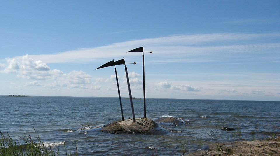 Bild på en skulptur på en kobbe i vattnet, det är tre stolpar med trekantiga flaggor i toppen.