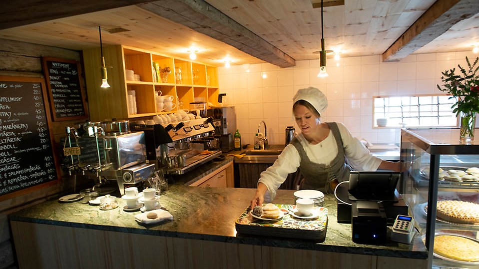 Kvinna som står bakom disken gör i ordning bricka med kaffe och kakor till gäst. Hon är tidsenlig klädd med snörliv över klänningen och huvudkläde.